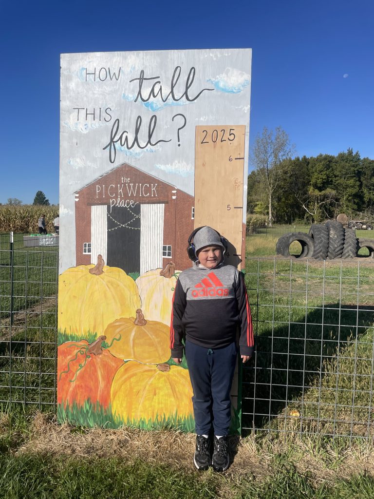 Student poses in front of the “How Tall This Fall” backdrop at The Pickwick Place during the High Road School of Bucyrus field trip, surrounded by colorful autumn decorations.