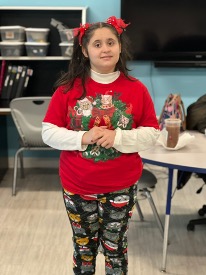 Kristina standing in a classroom smiling at the camera while wearing a bright red holiday shirt and matching festive hair bows. BEST Academy November student spotlight.