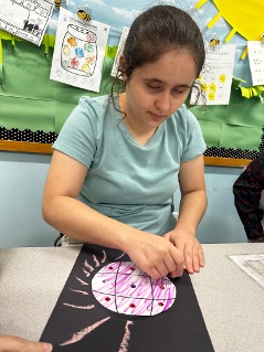 Kristina sitting at a classroom table working on a colorful solar system themed art project made with construction paper, chalk, and markers.