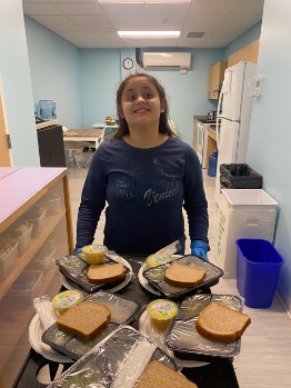 Kristina smiling in a lunch or break room with several prepared meal trays in front of her, highlighting her participation in daily routines at BEST Academy.