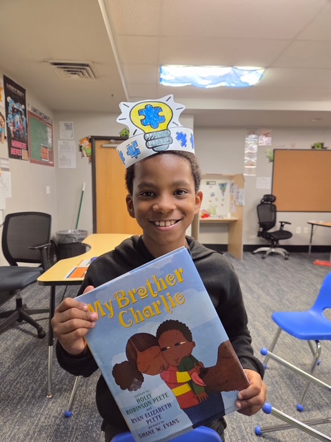 Un alumno con un gorro de papel que representa la «corona del pensamiento» sostiene el libro *Mi hermano Charlie* durante una clase de sensibilización sobre el autismo en la Sierra School del condado de Solano
