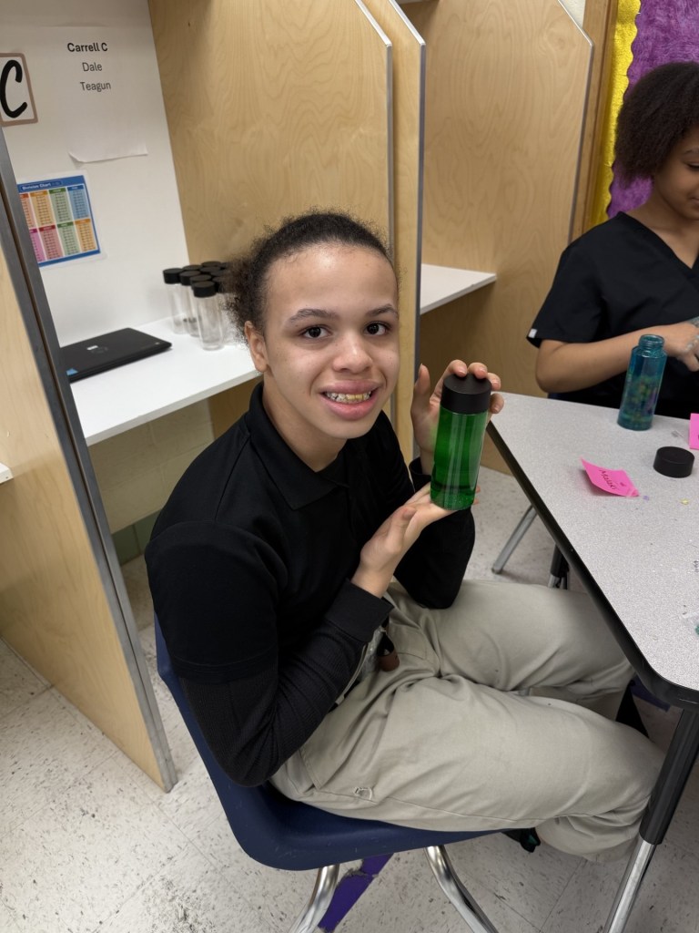 Un alumno sostiene una botella sensorial verde y sonríe durante un proyecto de apoyo al autismo