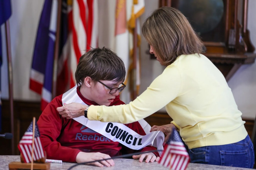 Cathy Payne, secretaria del Ayuntamiento de Wilkes-Barre, ayuda al miembro honorario del Ayuntamiento Jaxon Gillam a colocarse la banda durante un acto celebrado con motivo del Mes de Concienciación sobre el Autismo en el Ayuntamiento de Wilkes-Barre el viernes 10 de abril de 2026.