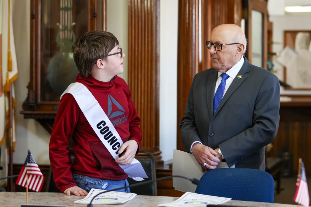 Cathy Payne, secretaria del Ayuntamiento de Wilkes-Barre, ayuda al miembro honorario del Ayuntamiento Jaxon Gillam a colocarse la banda durante un acto celebrado con motivo del Mes de Concienciación sobre el Autismo en el Ayuntamiento de Wilkes-Barre el viernes 10 de abril de 2026.