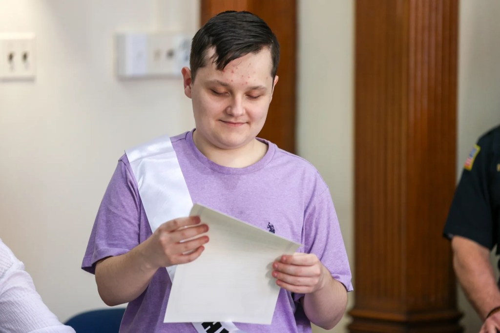 Cathy Payne, secretaria del Ayuntamiento de Wilkes-Barre, ayuda al miembro honorario del Ayuntamiento Jaxon Gillam a colocarse la banda durante un acto celebrado con motivo del Mes de Concienciación sobre el Autismo en el Ayuntamiento de Wilkes-Barre el viernes 10 de abril de 2026.