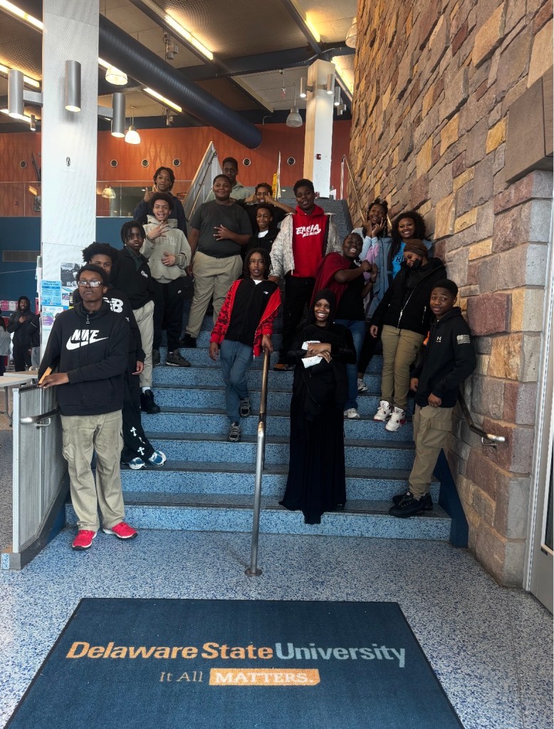 Los alumnos de la Excel Middle Years Academy posan en el interior de la Universidad Estatal de Delaware, junto a una escalera y una alfombra del campus, durante su visita a la universidad