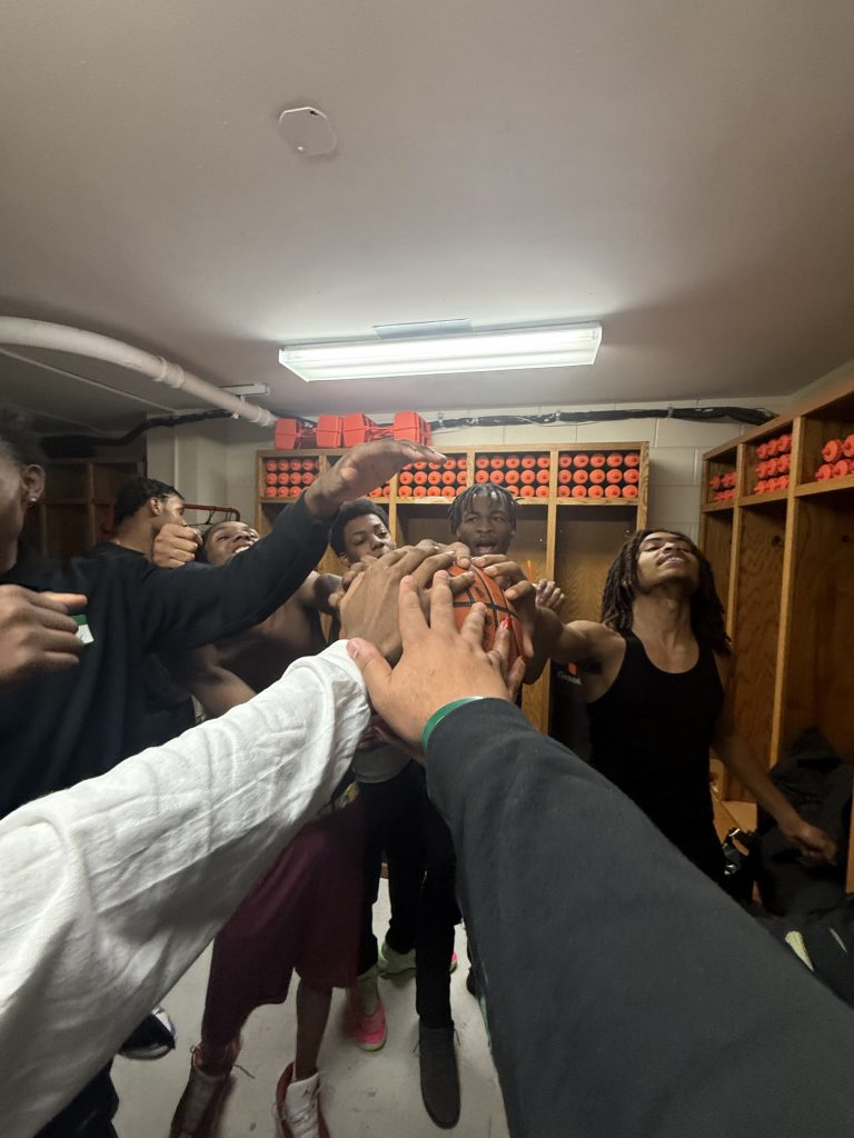 Excel Academy basketball team stands in a locker room huddle with hands raised before the game