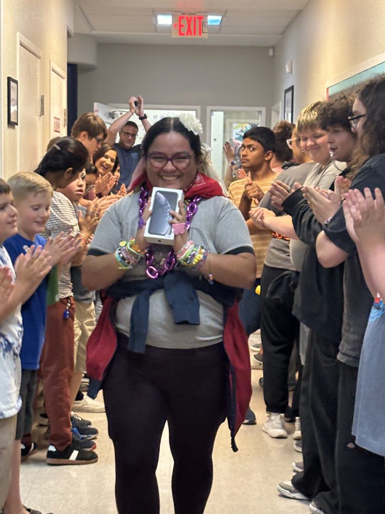 Yasmeen holding her iPhone while students and staff cheer in a hallway celebration at Sierra School of Gilbert CASE program
