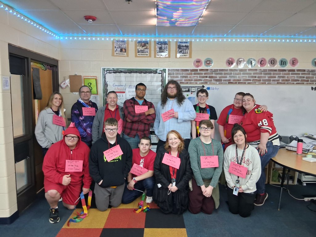 Un grupo de alumnos y personal posando en un aula con un cartel en el que se lee: «NWCA, estamos orgullosos de nuestro colegio».