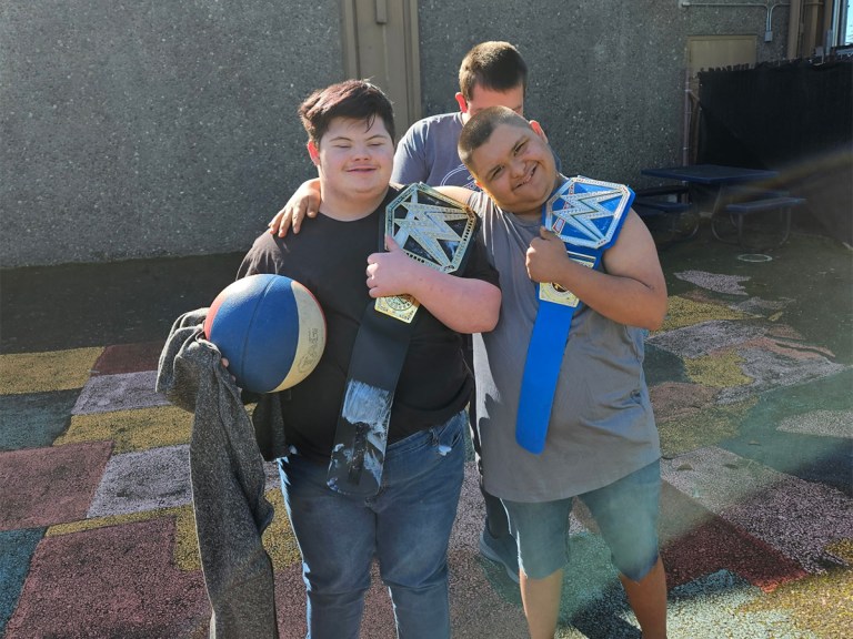 Students outdoors smiling with WWE championship belts over their shoulders while posing together with a basketball.