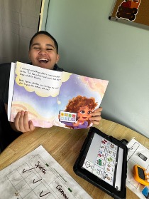 Estudiante sonriente, con un libro abierto en la mano, sentado en el pupitre de un aula con una tableta y material escolar