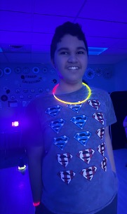 Un estudiante sonríe bajo la luz de neón, vestido con una camiseta de Superman y accesorios luminosos, durante una actividad escolar