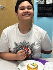 Un estudiante sonríe sentado en un pupitre, con un donut en un plato delante de él