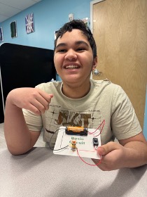 Un alumno sostiene con orgullo en el aula un proyecto de circuito básico con cables, conectores y una pila
