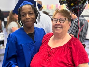 Valerie Quackenbush, miembro del personal de la High Road School de Germantown, sonríe junto a una alumna que se gradúa, vestida con toga y birrete.