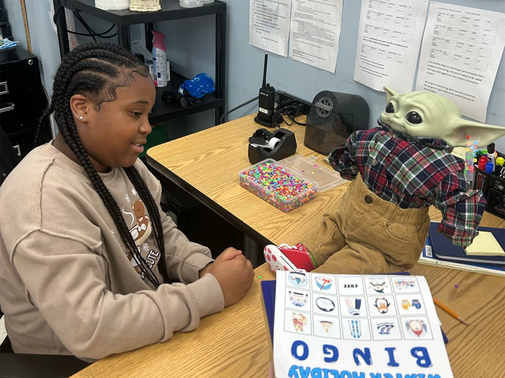 Un alumno sentado en el pupitre de un aula, con la mascota de Baby Yoda y las actividades de clase expuestas cerca.