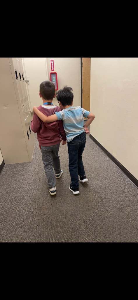 Two students walking down a school hallway near lockers with one student’s arm around the other.