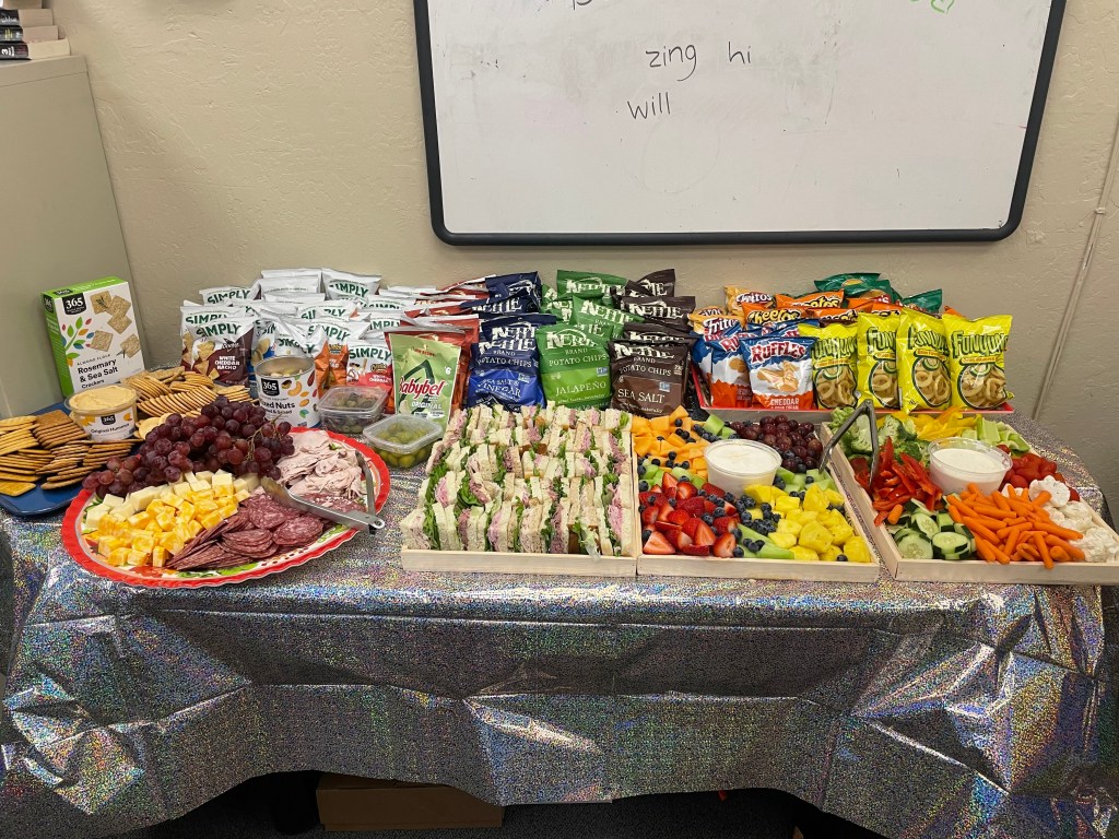 Table filled with snacks and lunch items including sandwiches, crackers, fruits, vegetables, chips, nuts, meats and cheeses.