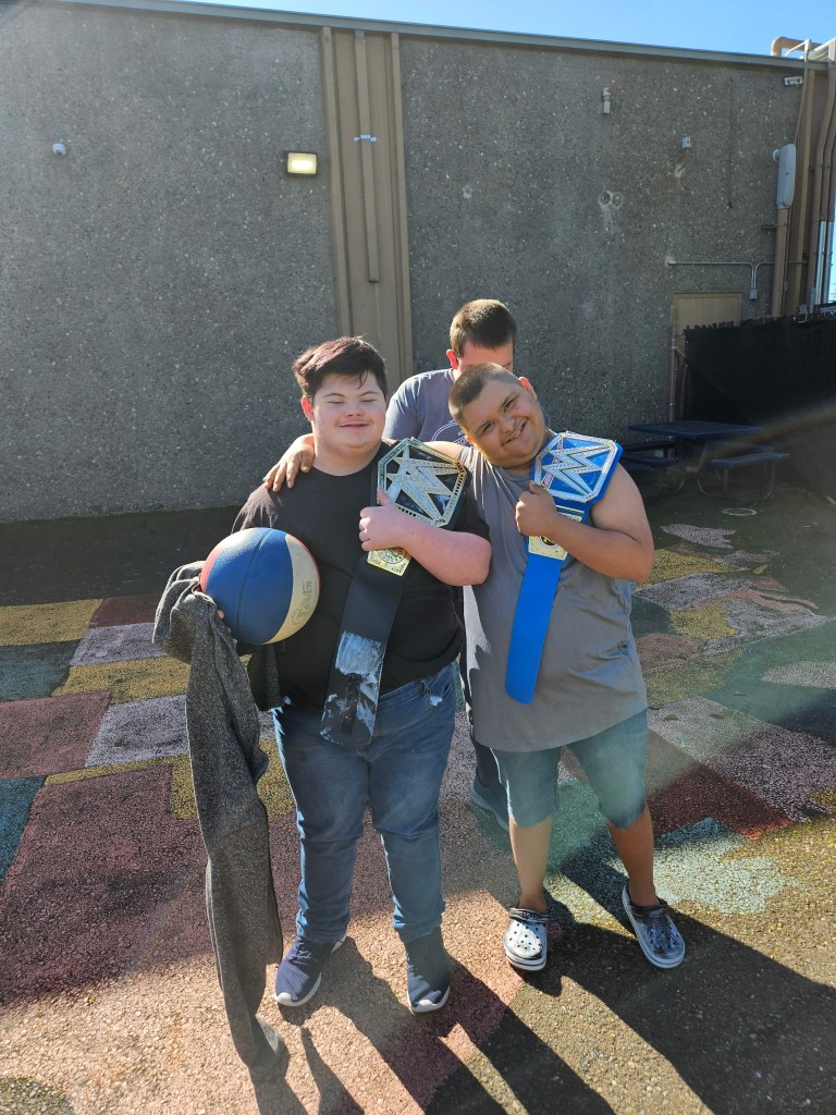 Students outdoors smiling with WWE championship belts over their shoulders while posing together with a basketball.