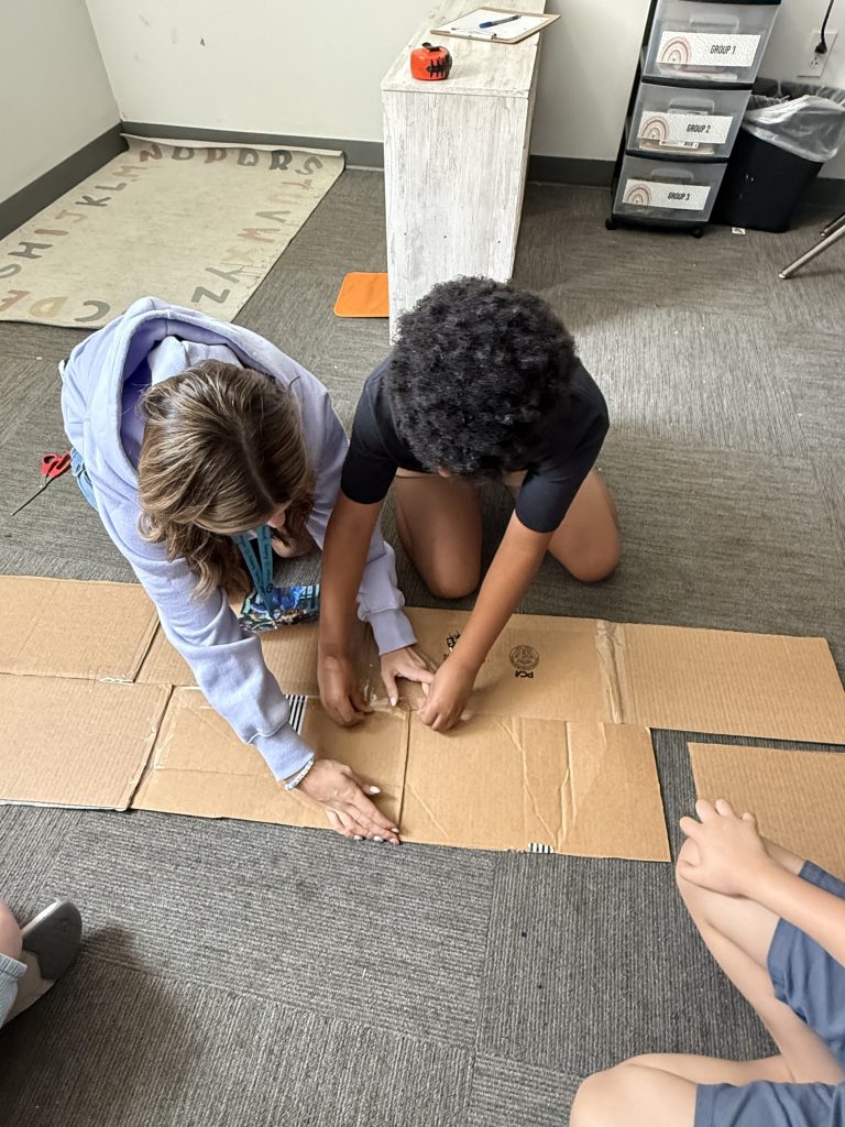 Teacher and student taping cardboard pieces together on the classroom floor during a hands-on learning activity.