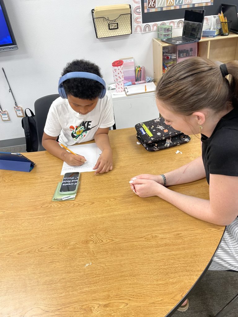 Student wearing headphones while writing at his desk as a staff member supports his work nearby.