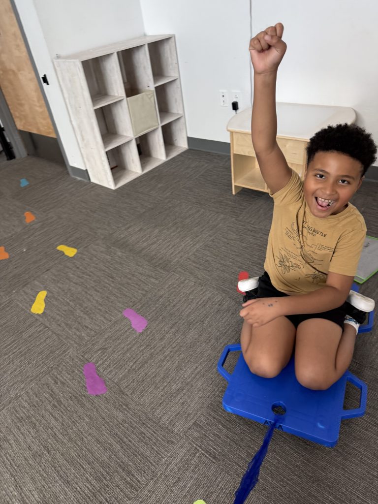Student kneeling on a classroom sled smiling at the camera with his fist raised in celebration.