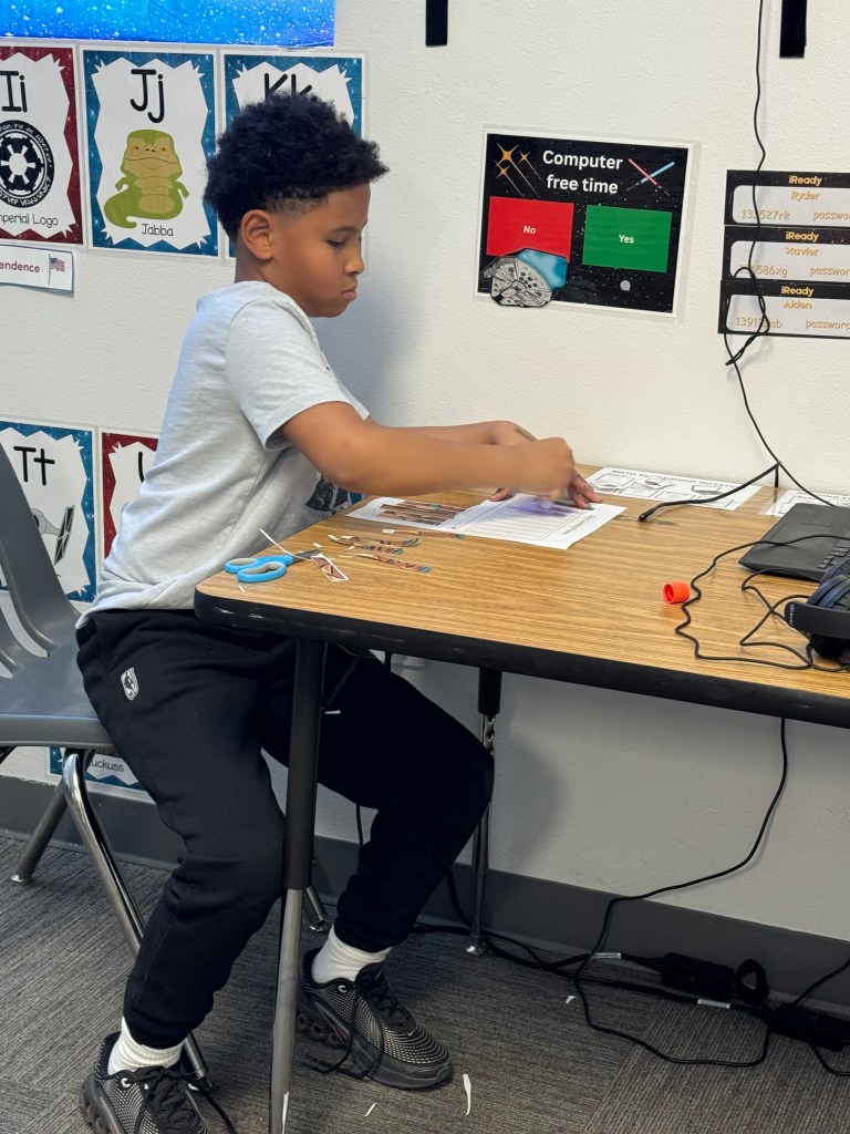 Student focused on a classroom paper and scissors project at his desk.