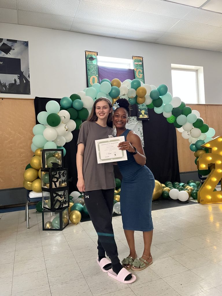Phoenix Academy graduates posing with certificates in front of a green, white and gold graduation balloon display during the Class of 2026 senior celebration