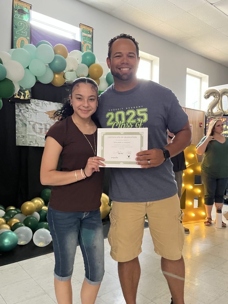 Phoenix Academy graduate smiling beside staff member while holding certificate of graduation during senior recognition event