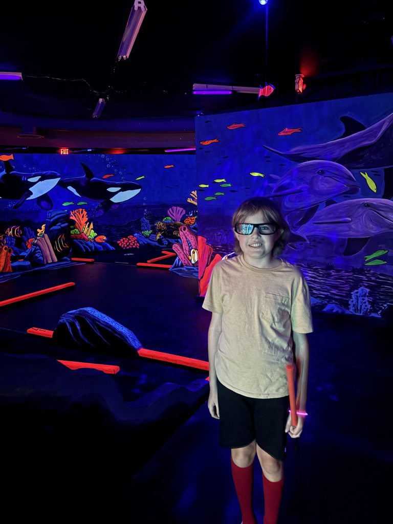 Student standing inside neon underwater-themed mini golf course during Sierra Gilbert CASE Fun Friday event.