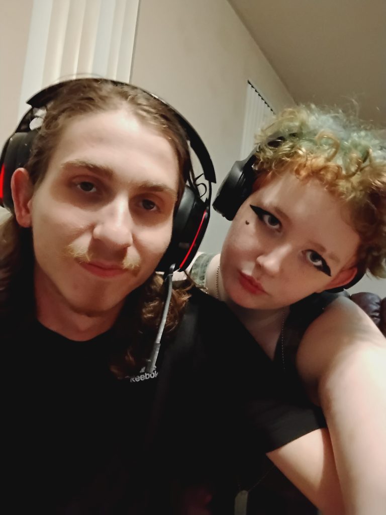 Tobi and a friend wearing headsets smile together while taking a selfie indoors.