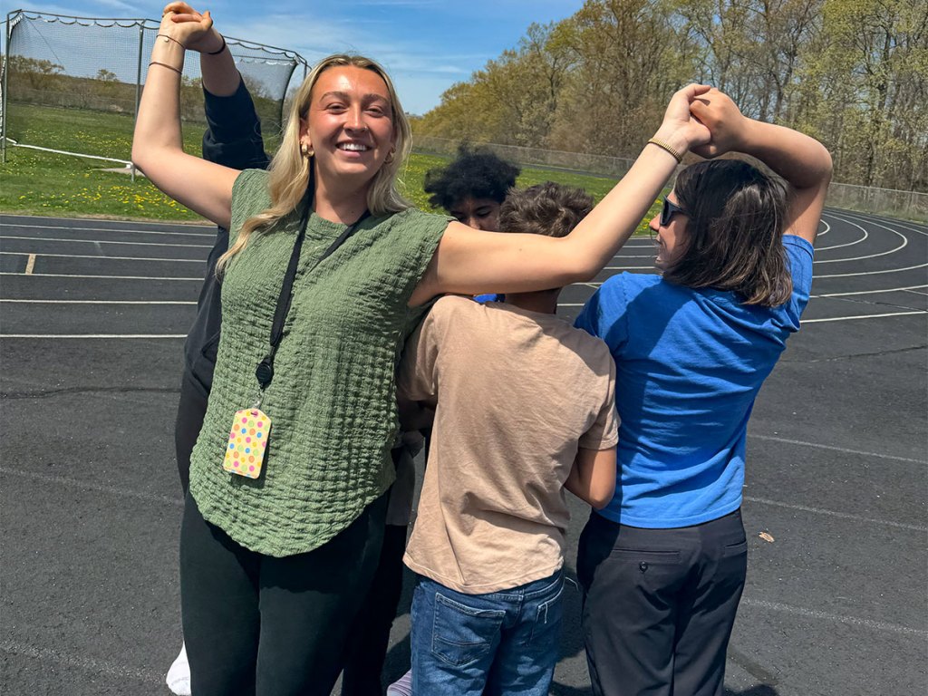 Staff member smiles while holding hands with students during an outdoor group activity near the school track.