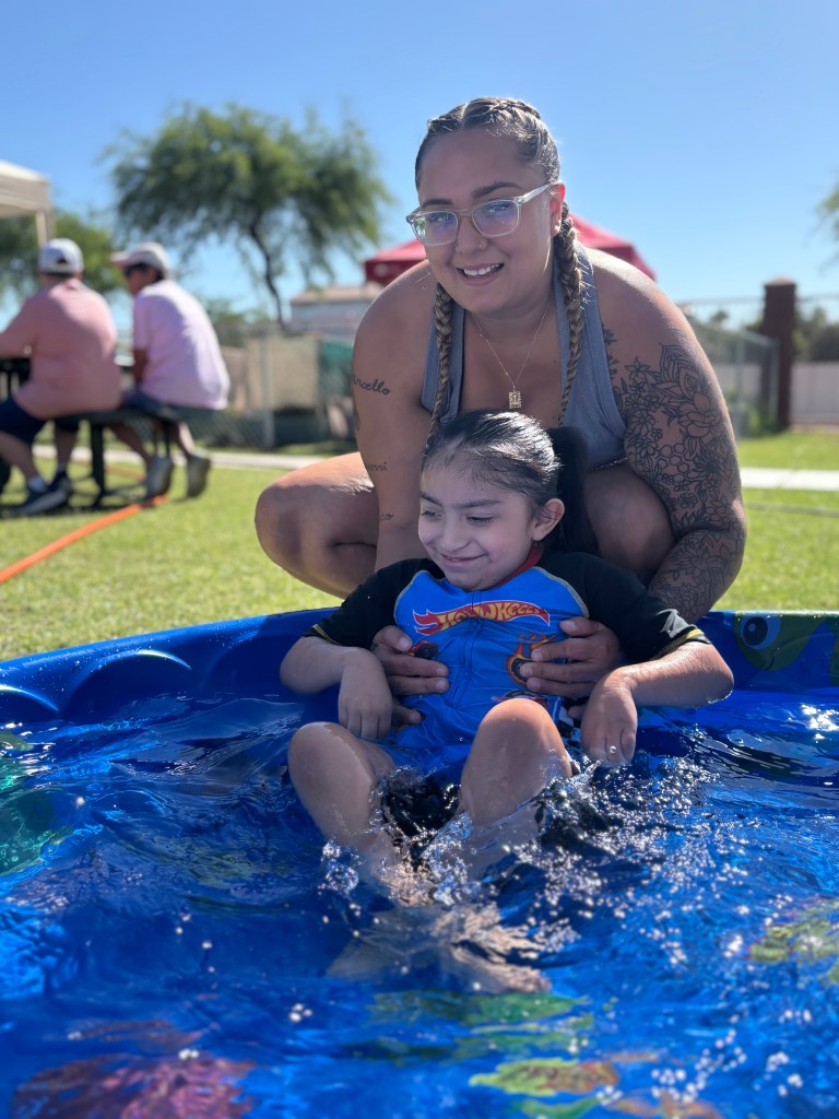 Un miembro del personal ayuda a un alumno sonriente a disfrutar de las actividades acuáticas en una piscina infantil durante la fiesta «Summer Splash Bash» de Sierra Gilbert.