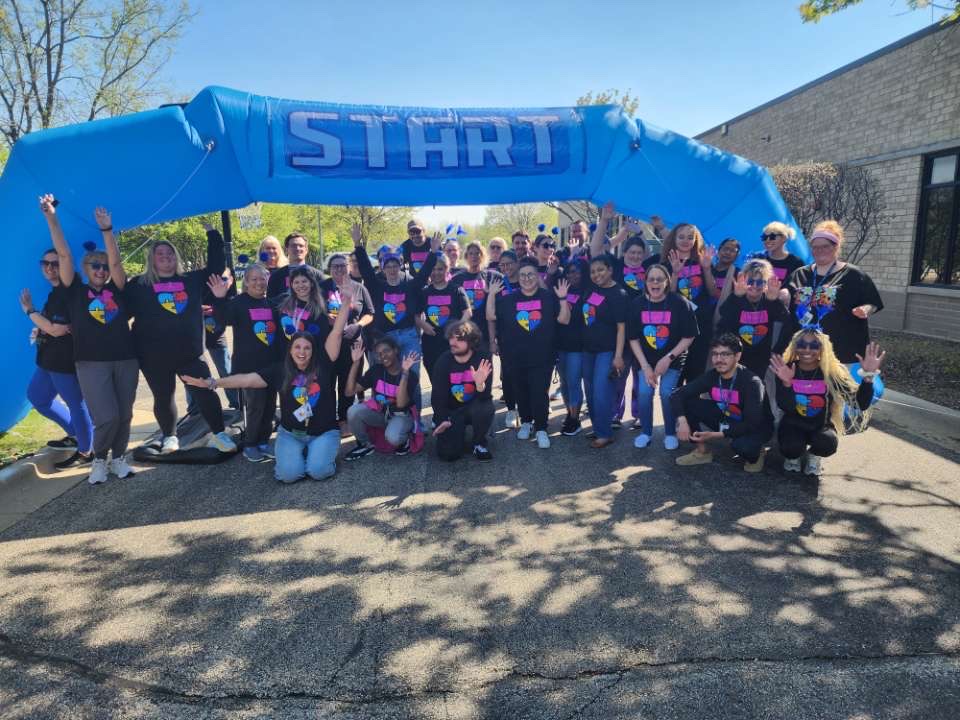 Students, staff, and community members posing together in matching autism awareness shirts near the Autism Walk starting line at High Road School of Hoffman Estates.