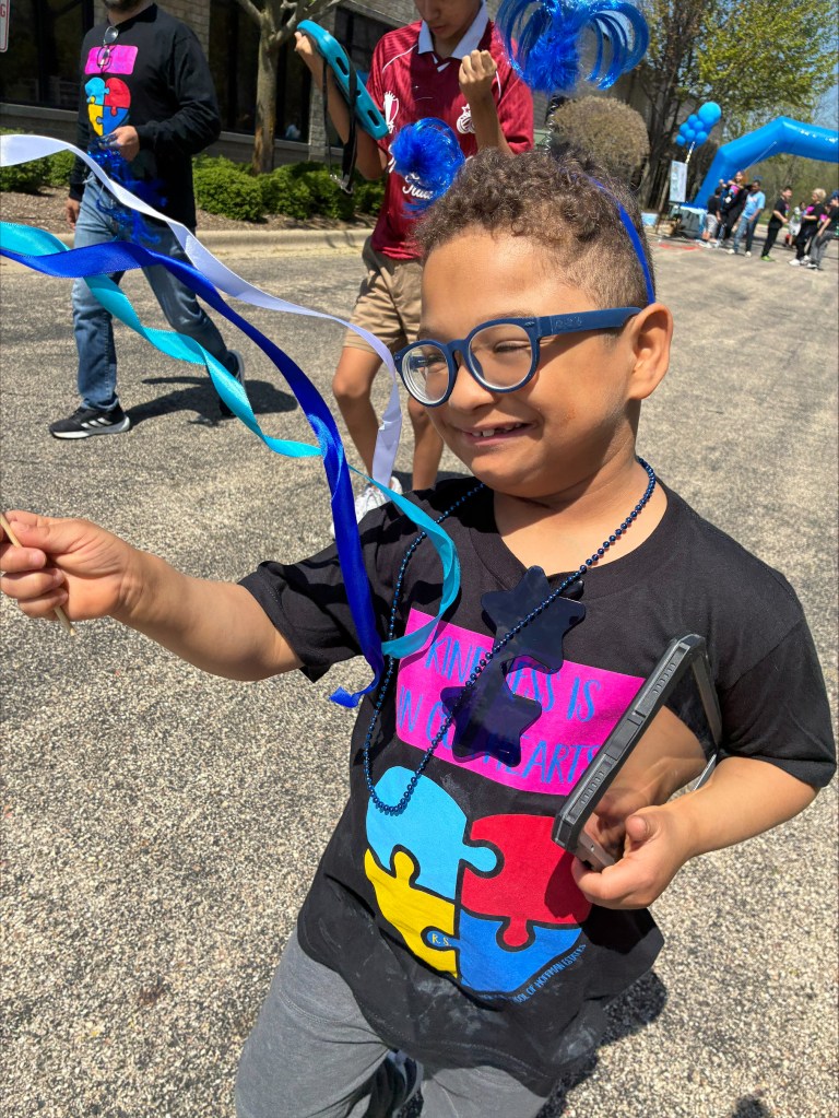Student smiling during the Autism Walk while wearing a decorated shirt, colorful accessories, and carrying a craft wand.