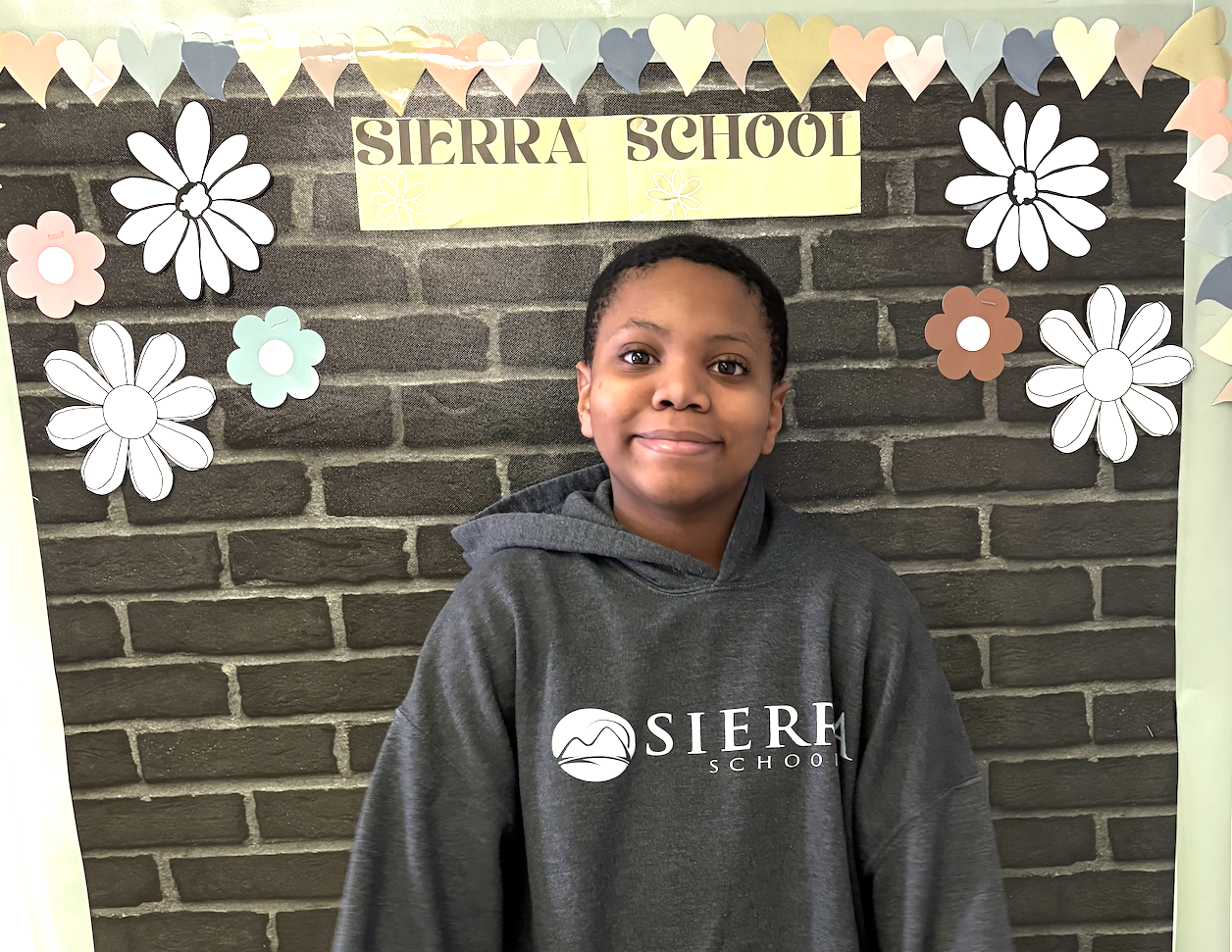 Student Josiah posing in front of a Sierra School display board decorated with flowers and hearts at Sierra School at McKinney