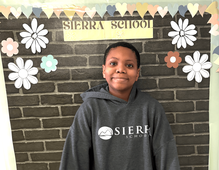 Student Josiah posing in front of a Sierra School display board decorated with flowers and hearts at Sierra School at McKinney