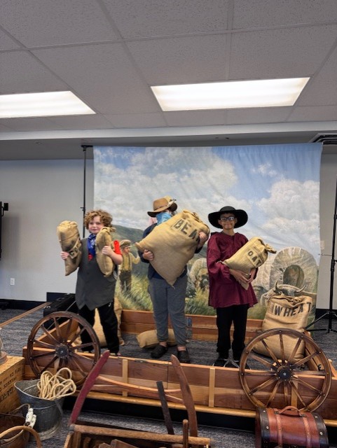 Students dressed in historical clothing holding labeled bags of oats, beans, wheat, and cans