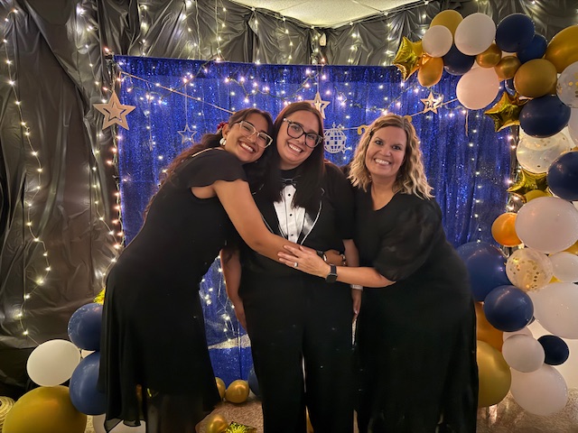 Staff posing in front of a decorated prom backdrop with balloons and lights