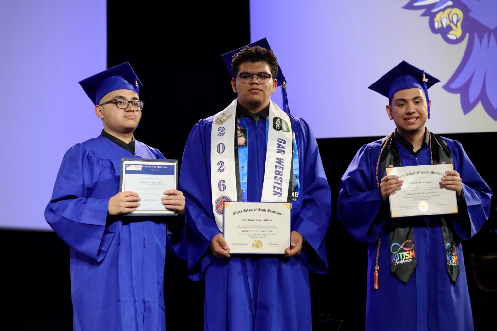 Three Sierra School of South Mountain graduates pose on stage in blue graduation gowns while holding diplomas during the Class of 2026 ceremony.