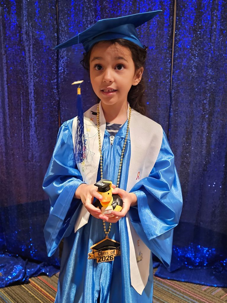 Kindergarten graduate Haiti J. smiles in a blue graduation cap and gown while holding a graduation keepsake at Sierra School of Scottsdale.