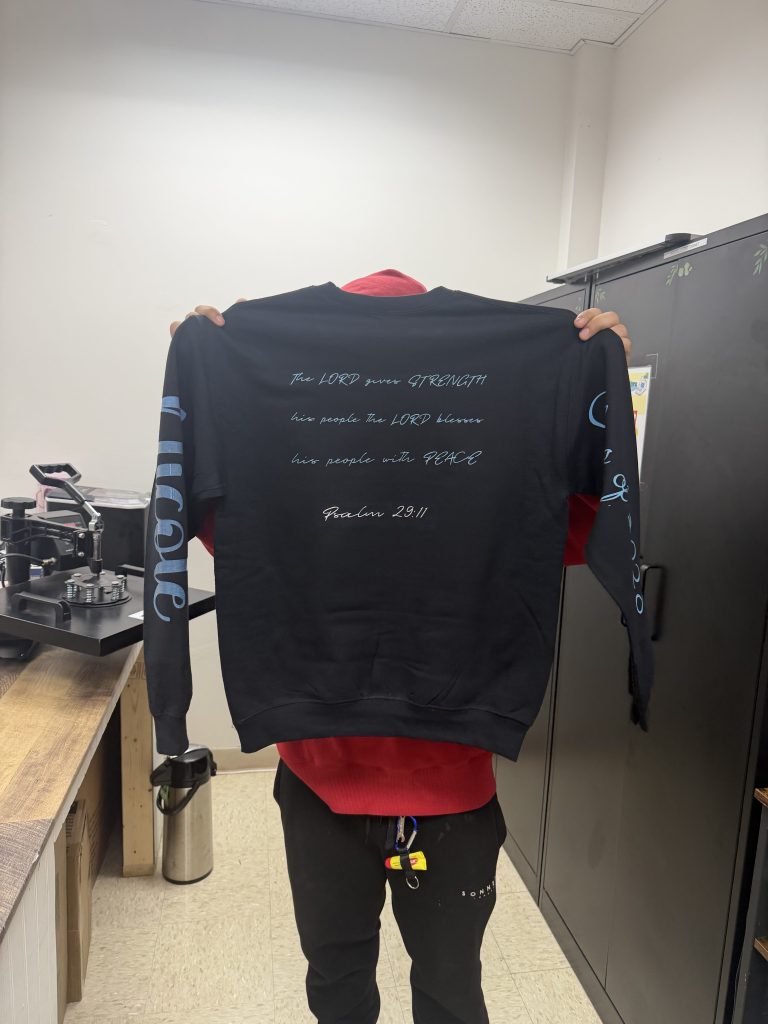 Un alumno muestra la parte trasera de una sudadera negra personalizada con un versículo bíblico, creada durante un proyecto artístico basado en la tecnología.