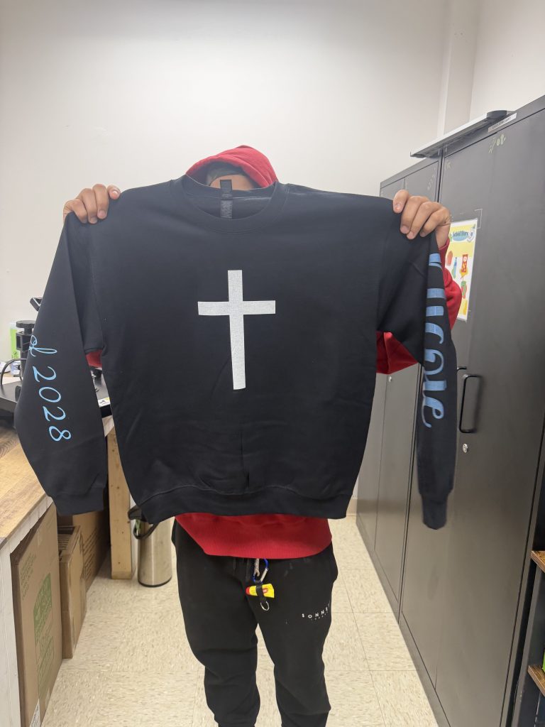 Un estudiante sostiene una sudadera negra personalizada con un diseño de cruz, creada con un programa de diseño digital, una máquina Cricut y una prensa térmica.
