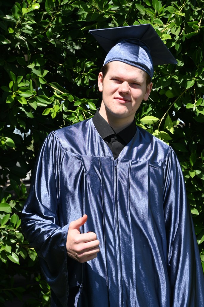 Greg poses outdoors in a graduation cap and gown and gives a thumbs-up after completing his studies at Sierra School of Peoria.