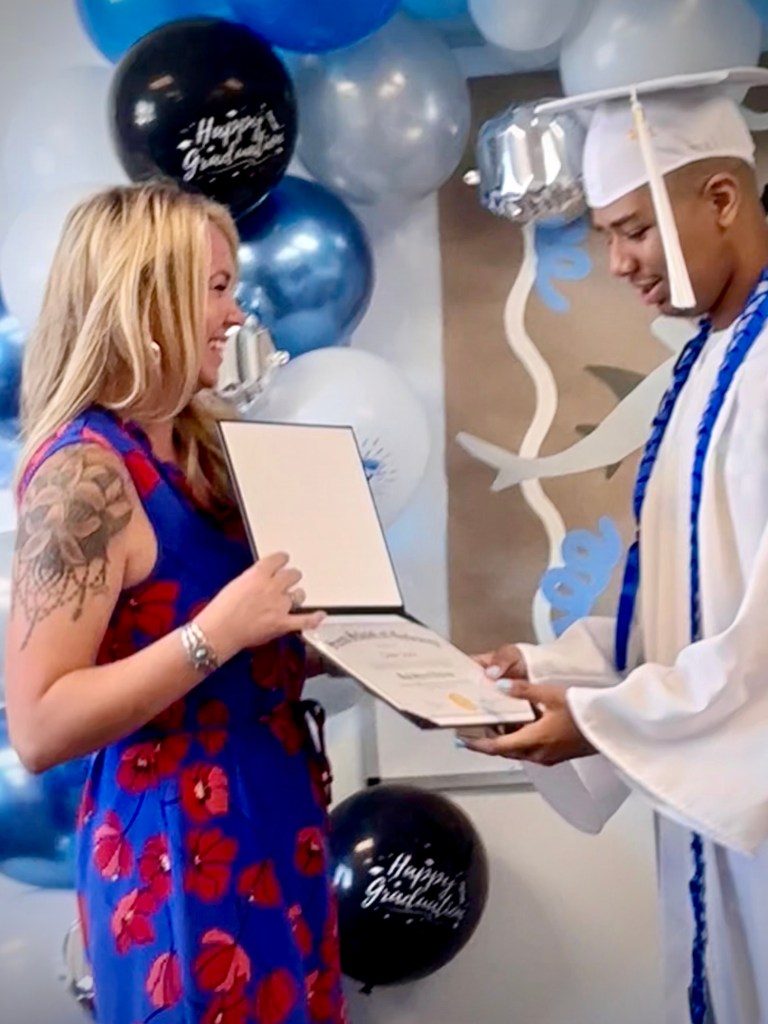 A graduate accepts a diploma during Sierra School of Gateway's graduation ceremony surrounded by blue and white decorations.