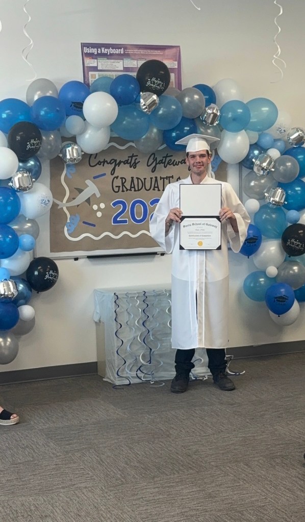 A Sierra School of Gateway graduate proudly displays a certificate in front of a Class of 2026 graduation backdrop.