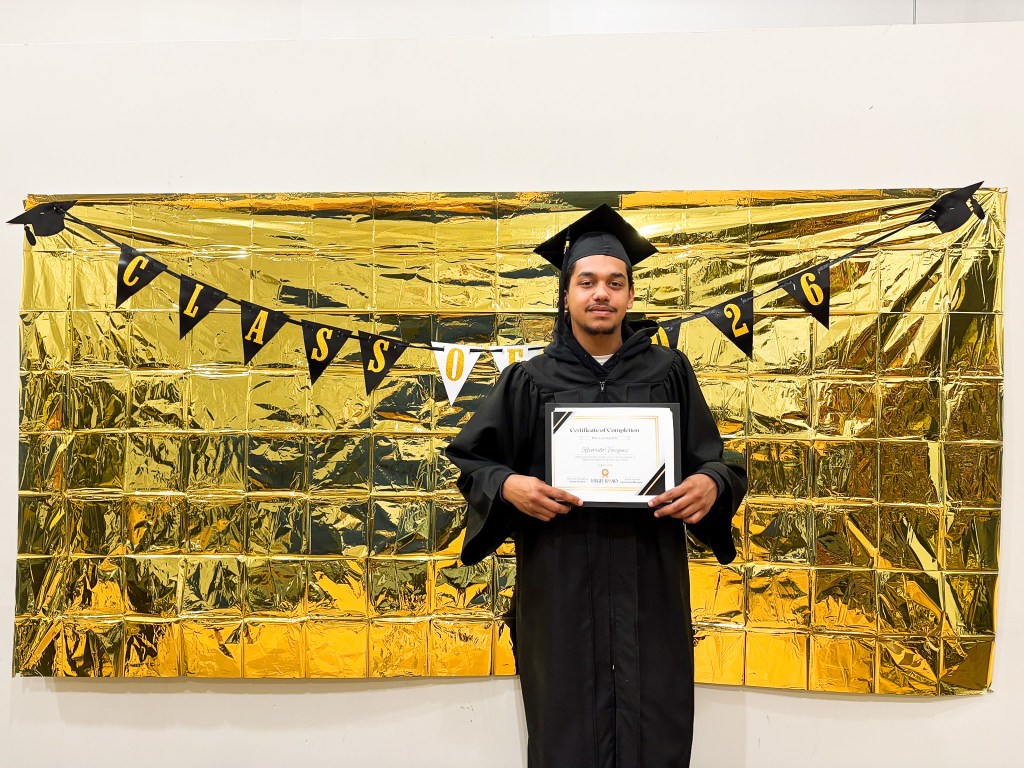 Un graduado de la High Road School de Hartford posa con un certificado delante de un telón de fondo de la promoción de 2026.
