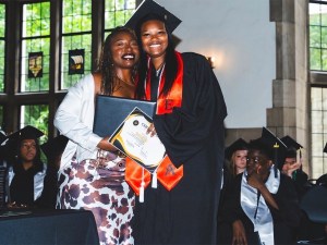 Una graduada de la Excel Middle Years Academy sonríe con toga y birrete mientras sostiene su certificado durante una ceremonia de reconocimiento de octavo curso.
