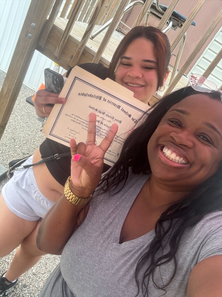Un graduado y un miembro del personal se hacen una selfie para celebrar el momento, sosteniendo un título del Distrito Escolar de Filadelfia.