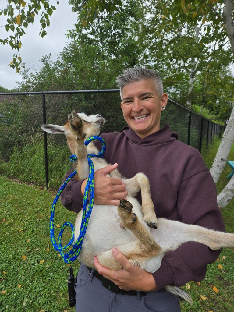 Un miembro del personal, con una sonrisa, sostiene a una de las cabras de la Graham Academy al aire libre.